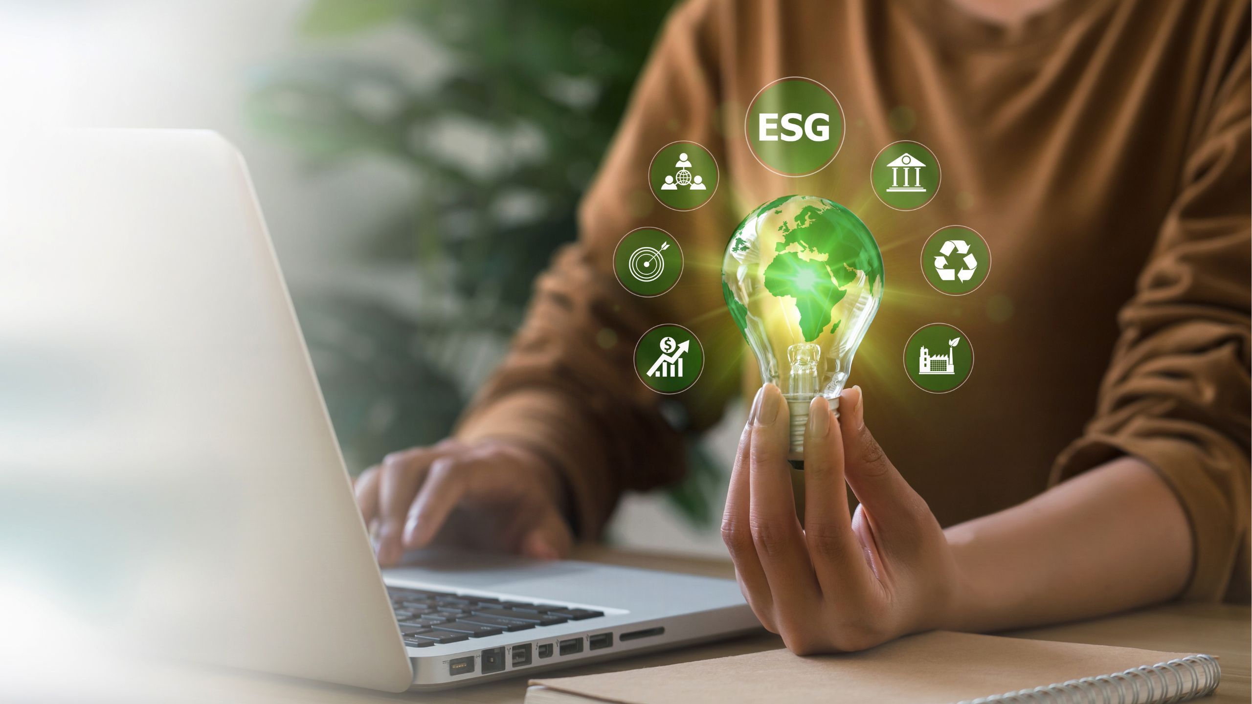 An person works in front of a laptop and holds a lit light bulb with icons representing sustainable business practices